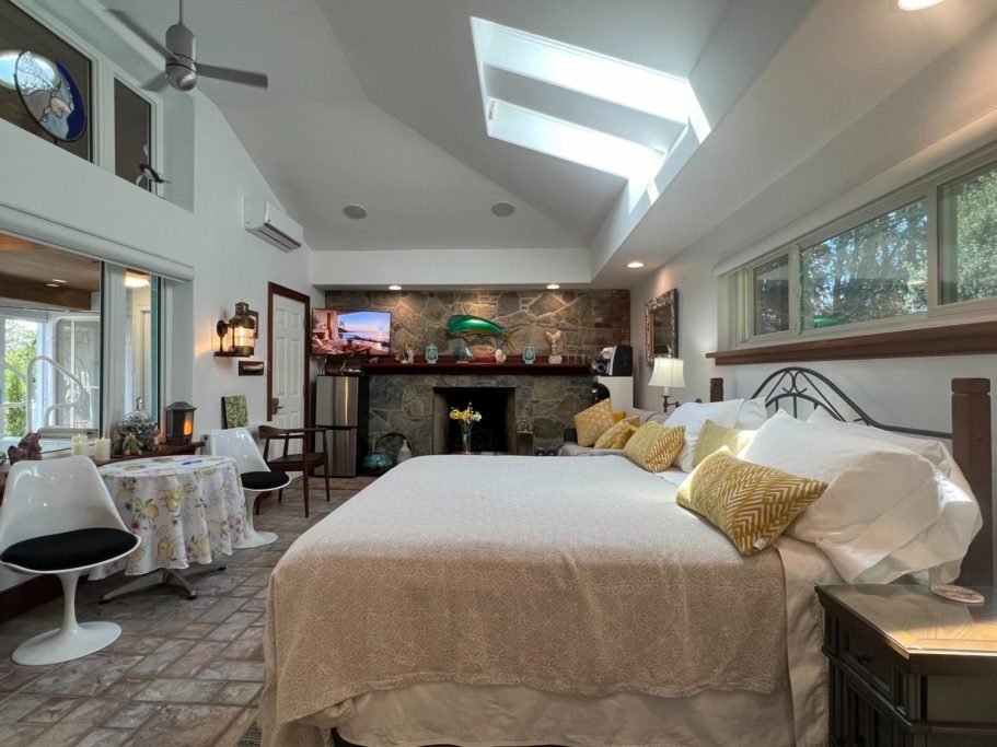 Wide panoramic view of the luxury guest suite showing the open layout, breakfast dining table, king bed, and stone fireplace at Wind in the 