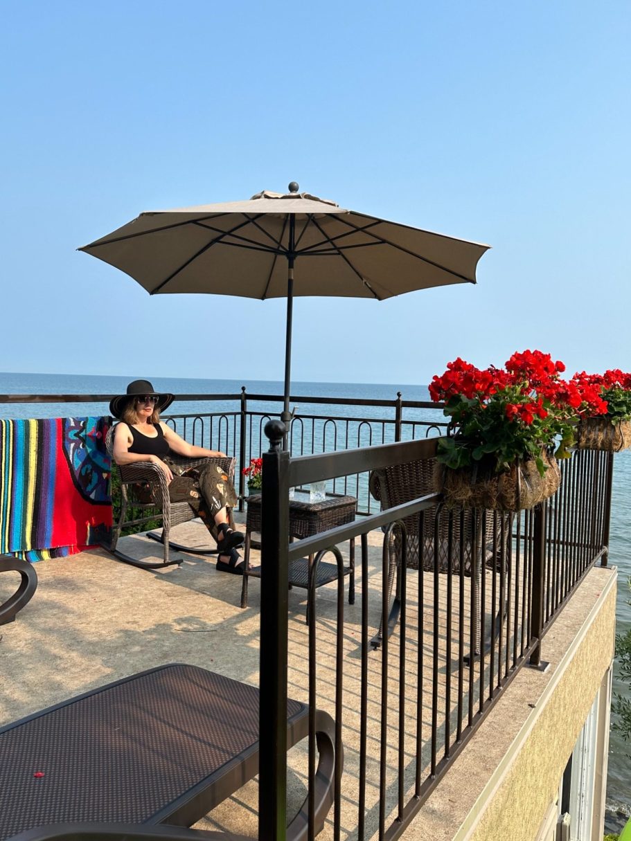 Lakeside patio with lounge chairs, umbrella, bright flowers, and panoramic views of Lake Ontario at Wind in the Willows Oasis.